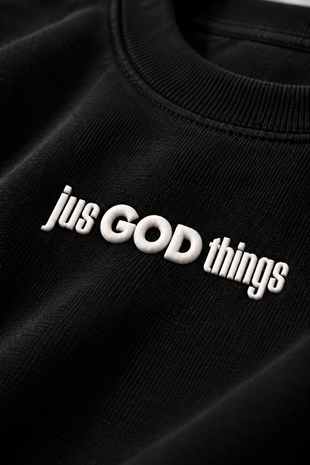 Close-up of the white Jus God Things raised puff-print logo on the black sweatshirt fabric.