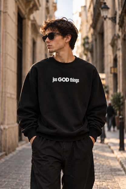 Male model wearing a black heavyweight JGT oversized sweatshirt with white chest branding, styled with black trousers in an urban setting.