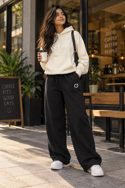 Female model wearing black JGT relaxed-fit sweatpants with a cream sweatshirt in an urban streetwear setting.